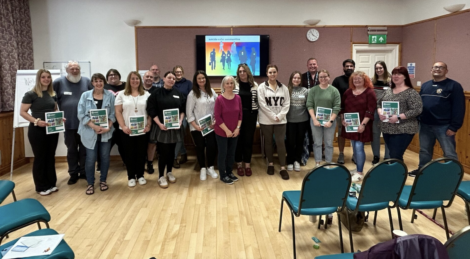A group of people standing in a room holding certificates, with chairs and a presentation screen visible in the background.