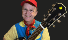 A person wearing a red cap, glasses, and a blue and yellow outfit is smiling and holding a guitar.