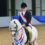 A person in a riding uniform and helmet smiles while seated on a white horse adorned with red, white, and blue ribbons in an indoor arena.