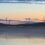 Landscape at dawn with several wind turbines situated on hills. Calm waters in the foreground reflect the turbines and the pastel-colored sky.