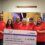 A group of six people in matching red shirts hold a large donation check for £227,538.69 to Cancer Research UK, standing inside Hay's Dock Café Restaurant.