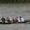 A rowing team of eight people is paddling in a boat with a blue and white flag on a river.