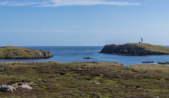 A panoramic view of a coastal landscape with grassy terrain, rocky hills, a lighthouse on an island, and several wind turbines visible on the horizon under a clear blue sky.