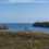 A panoramic view of a coastal landscape with grassy terrain, rocky hills, a lighthouse on an island, and several wind turbines visible on the horizon under a clear blue sky.