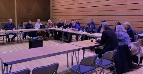 A group of people are seated around a U-shaped arrangement of tables in a room with wooden walls. Some participants are gesturing while others listen attentively.