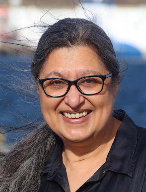 A person with long, dark hair, glasses, and a black shirt smiles in front of a body of water.