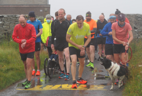 A group of runners and a person in a wheelchair gather at a wet starting line on a foggy day. A dog on a leash is barking or excited.