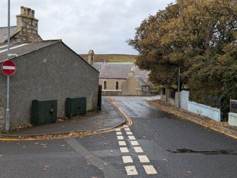 Lightly wet road with a no-entry sign, houses with stone walls, and autumn trees on both sides. A distant view of grassy fields under a cloudy sky in the background.
