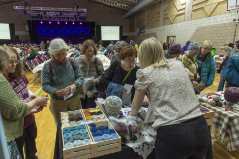 People browse and purchase items from a vendor's table at a craft fair inside a gymnasium, with a stage and seating area in the background.