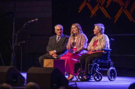 Three people are seated on a stage, with one person using a wheelchair. A woman in the center holds a microphone.