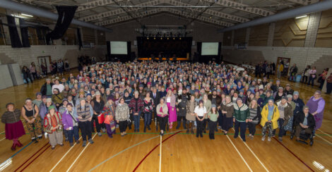 A large group of people standing in a gymnasium, facing the camera. In the background, there is a stage with a banner and a few people seated. Athletic markings are visible on the floor.