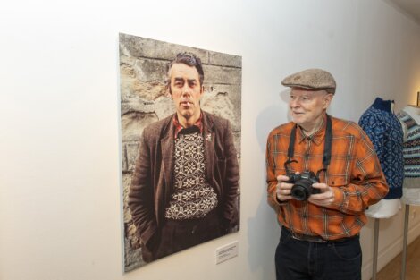 An elderly man wearing an orange plaid shirt and flat cap holds a camera, standing next to a large portrait of another man displayed on a museum or gallery wall.