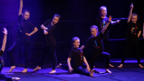 Group of young dancers in black outfits performing a choreographed routine on stage with dramatic lighting.