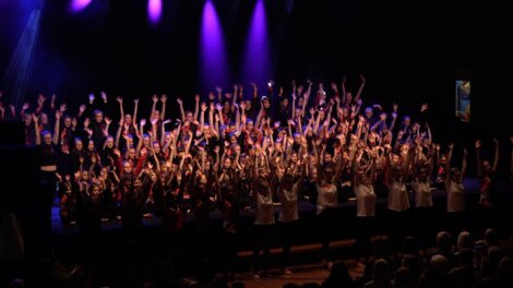 A large group of people on stage with their arms raised, illuminated by purple stage lights, performing in a dark auditorium.