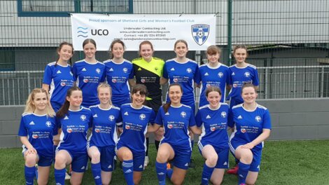 A women's football team poses for a group photo in blue uniforms in front of a sponsorship banner.
