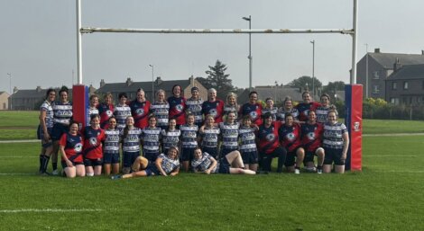 A group photo of two female rugby teams posing under a goalpost on a grassy field with housing in the background.