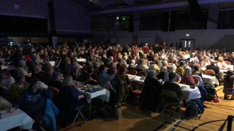 A large audience sits at tables in a dimly lit auditorium, attentively facing a stage not visible in the image.
