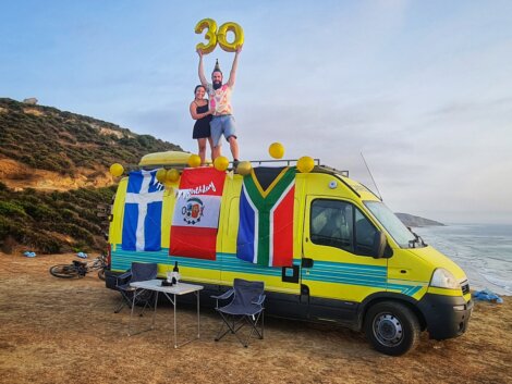 Two people on top of a yellow van holding balloons shaped like "30" with various flags on the van, two camping chairs and a table set up nearby, located on a coastal cliffside.