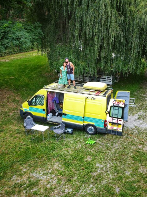 Two people stand on the roof of a yellow camper van parked on grass. Chairs and camping equipment are set up beside the van. Large trees and greenery are in the background.