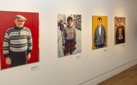 Four portrait photographs of people wearing patterned sweaters are displayed on a white gallery wall. The individuals vary in age and background, and each photo includes a small label beneath it.