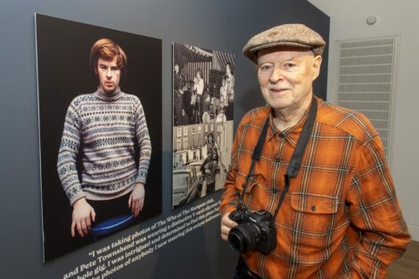 An elderly man wearing a plaid shirt and flat cap holds a camera while standing beside a display featuring photographs of various subjects, including one in a patterned sweater.