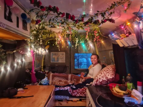 A person smiling while sitting inside a decorated van with floral garlands and colorful string lights. The van interior includes maps, storage shelves, a bed with a blanket, and a small kitchen area.