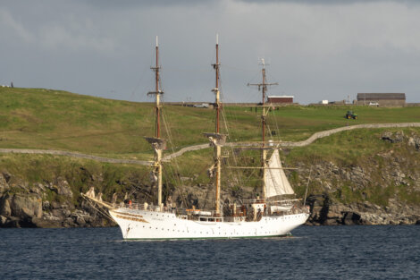 A tall ship with multiple masts and sails is anchored near a rocky shoreline with grassy hills and a few buildings in the background.