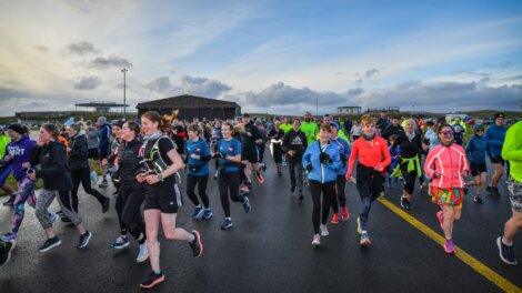 A large group of people in athletic gear participate in an outdoor run on a road under a cloudy sky.