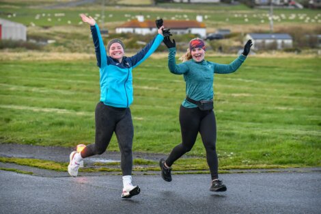 Two people in athletic clothing jog on a paved path, smiling and raising their arms exuberantly. A grassy field and distant buildings are in the background.