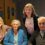Four people sitting together at a table, with three smiling at the camera. The group consists of two older individuals and two younger women. They are indoors, in a room with grey walls and wooden accents.
