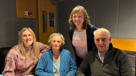 Four people sitting together at a table, with three smiling at the camera. The group consists of two older individuals and two younger women. They are indoors, in a room with grey walls and wooden accents.