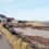 A narrow road winds past a white house and stone buildings by a rocky coastline with sparse vegetation and a small sandy beach. The sky is clear with light clouds.