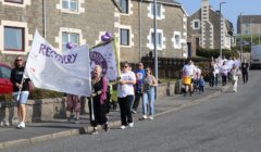 A group of people participate in a community march, carrying banners and balloons, walking down a residential street in daylight.