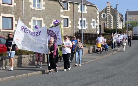 A group of people participate in a community march, carrying banners and balloons, walking down a residential street in daylight.