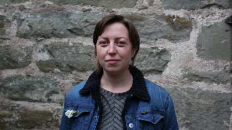 A person with short hair stands in front of a stone wall, wearing a denim jacket with a white flower pinned to it.