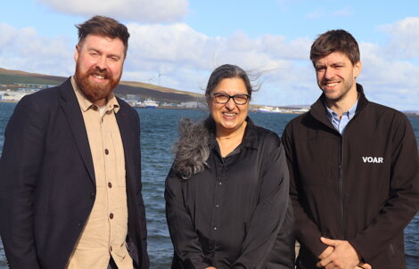 Three people standing outdoors by a body of water with a wind turbine and industrial buildings in the background.