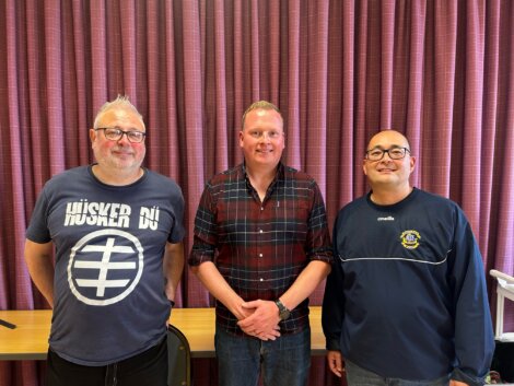 Three men standing side by side indoors. Each is wearing casual clothing. A table and chair are visible behind them, and a curtain with a vertical striped pattern forms the backdrop.