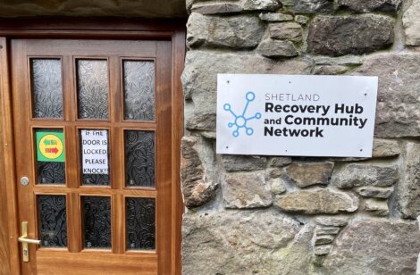 A wooden door next to a stone wall with a sign that reads "Shetland Recovery Hub and Community Network". The door has a smaller sign stating "IF THE DOOR IS LOCKED PLEASE KNOCK!.