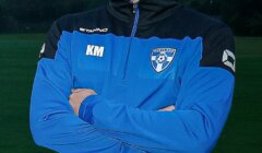 Four individuals standing outside with arms crossed, wearing blue and black jackets with team logos, and smiling for the camera. A grassy field is in the background.
