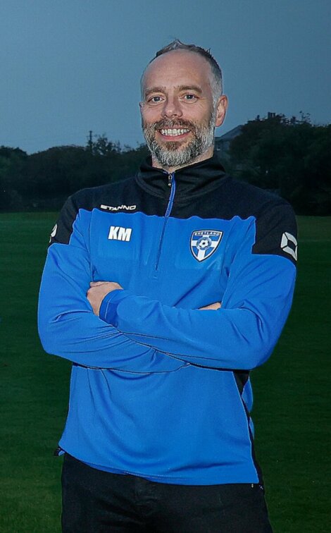 Four individuals standing outside with arms crossed, wearing blue and black jackets with team logos, and smiling for the camera. A grassy field is in the background.