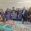 A group of people, some in aprons and hair nets, pose in front of a table filled with containers of variously colored liquids in a laboratory or production setting.