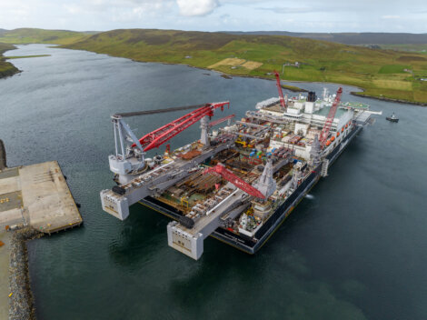 A large offshore construction vessel with cranes docked in a calm body of water near a hilly landscape.