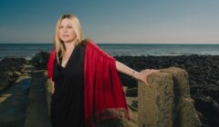 A woman with long blonde hair, wearing a black dress and red shawl, stands on a rocky coastline with her hand on a cement structure, with the sea and a clear sky in the background.