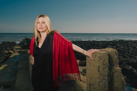 A woman with long blonde hair, wearing a black dress and red shawl, stands on a rocky coastline with her hand on a cement structure, with the sea and a clear sky in the background.