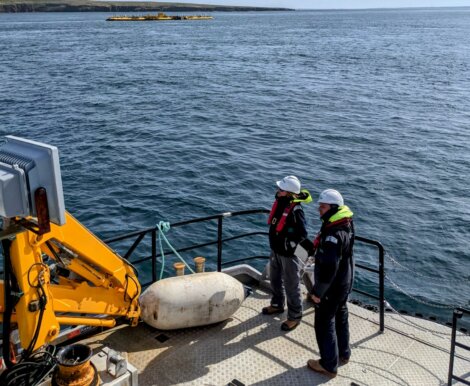 Two people in safety gear stand on a boat's deck, monitoring equipment. An island is seen in the distance across the water.