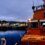 An orange RNLI lifeboat docked at a harbor with town buildings and evening lights in the background under a cloudy sky.