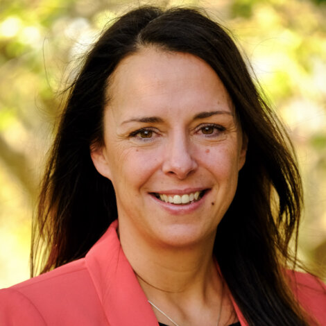 A woman with long dark hair smiles, dressed in a coral jacket outdoors with a leafy background.