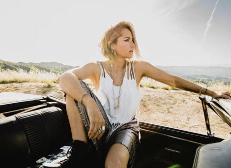 A person with short hair sits in a convertible car, looking into the distance. They are wearing a white tank top and shorts. The background features a sunny landscape with hills and vegetation.
