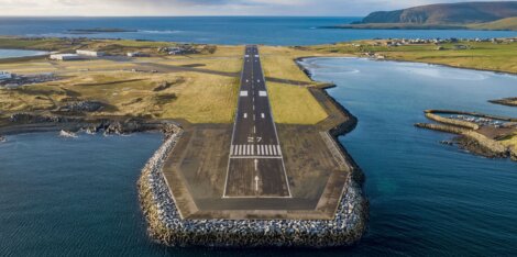 Aerial view of a coastal runway extending into the sea with adjacent green fields and small buildings.