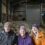 Three people, two elderly and one younger, wearing winter clothing and knit caps, pose inside a barn with sheep in the background.